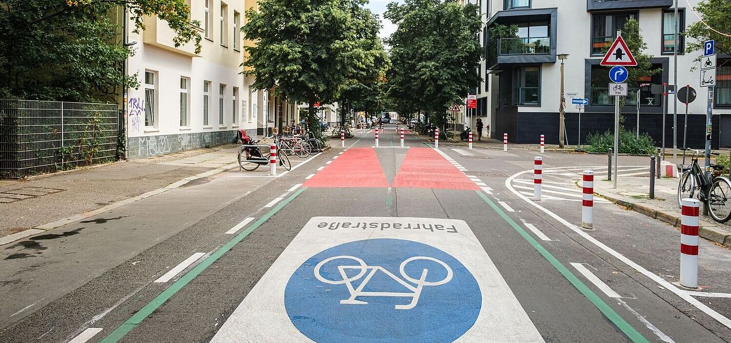 Eine Straße in Berlin: Rechts und links sind Häuserzeilen zu sehen, die Fahrbahn ist mit einem großen Fahrradsymbol versehen. Weiter hinten ist die Fahrbahn rot eingefärbt, Poller dienen als modaler Filter.  